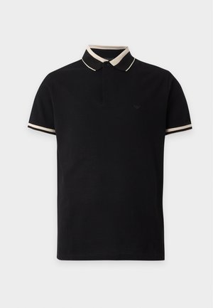 Bărbat care poartă un tricou polo negru cu tiv alb, blugi albastru închis, adidași negri și ochelari de soare negri, stând cu o mână în buzunar.