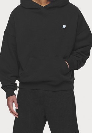 Person trägt einen schwarzen Hoodie mit einem kleinen blauen und weißen "P"-Logo auf der Brust, eine Hand in der vorderen Tasche und schwarze Hose.