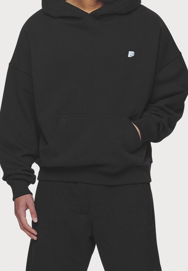 Personne portant un sweat à capuche noir avec un petit logo "P" bleu et blanc sur la poitrine, une main dans la poche avant et un pantalon noir.