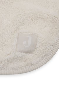 Crème-kleurig pluche dekentje met een zachte, donzige textuur en een genaaid label met een witte letter "J". Ronde rand met beige afwerking.