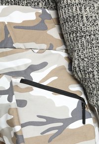 Beige och grå kamouflage cargobyxor med en zipficka. Tyget ser slätt ut, tillsammans med en texturerad grå stickad topp som delvis är synlig.