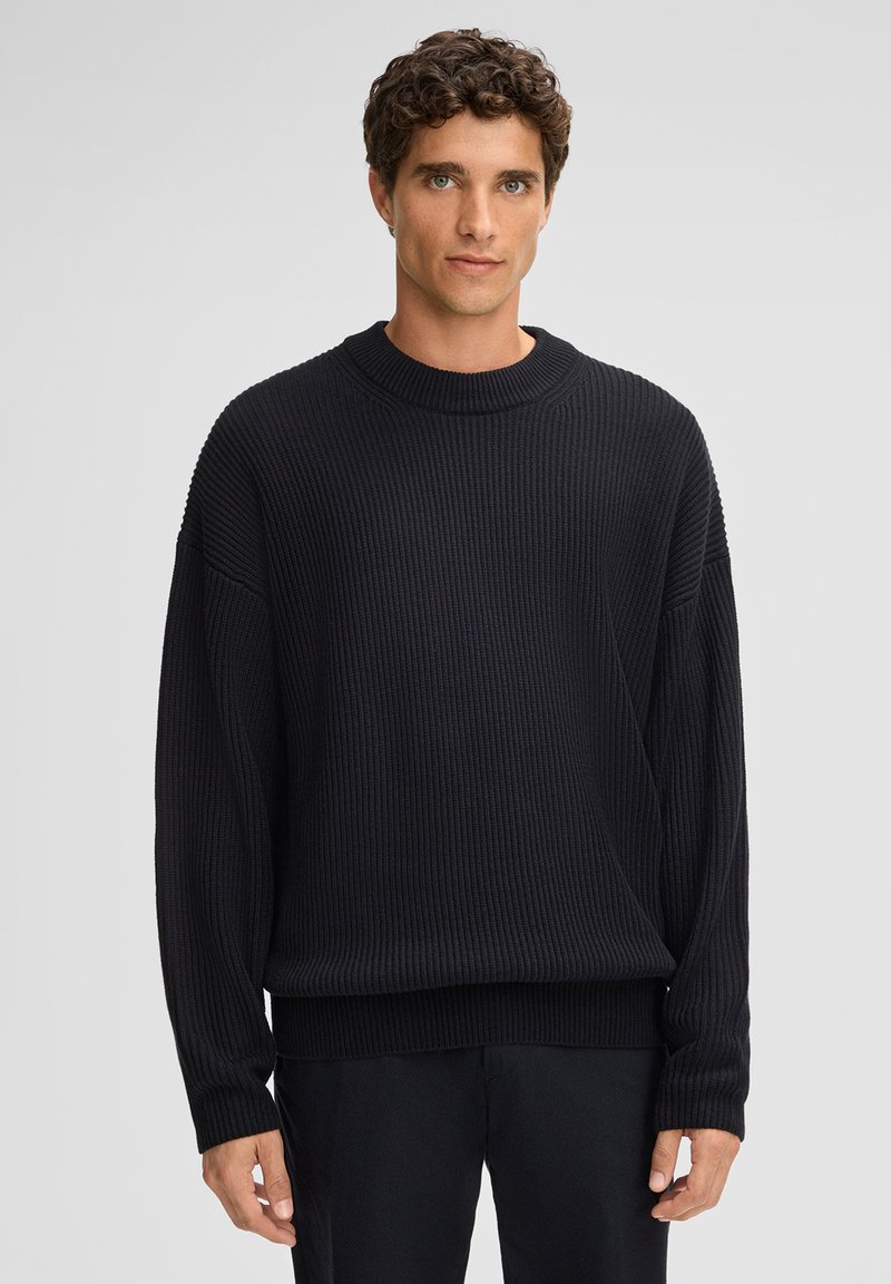 Giovane uomo con capelli ricci, che indossa un ampio maglione nero a coste e pantaloni neri, in piedi contro uno sfondo grigio chiaro uniforme.