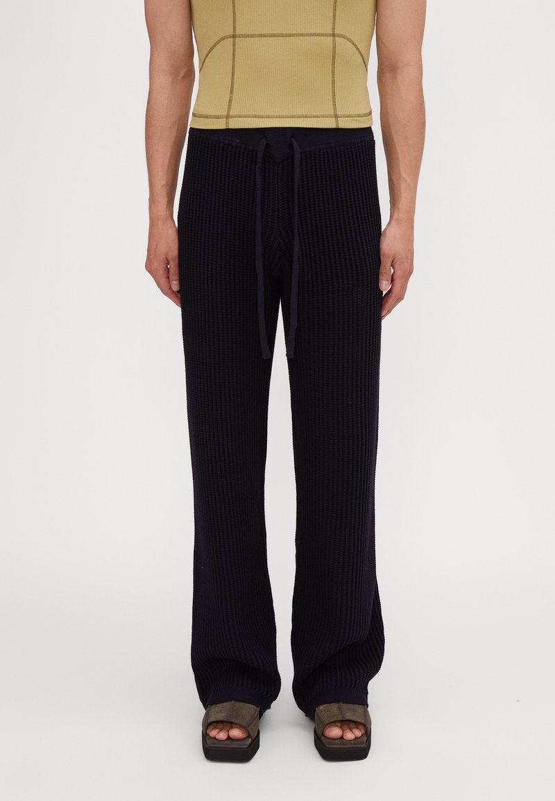Pantaloni tricotati în dungi de culoare bleumarin, cu o croială lejeră, talie cu şnur și picioare late, asortați cu sandale cu platformă maro.
