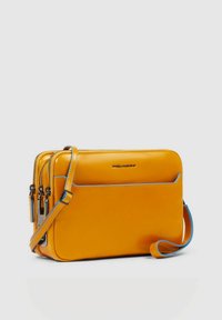 Geel leren crossbody tas met drie ritsvakken, blauwe bies en een verstelbare riem. Soepele textuur en rechthoekige vorm.