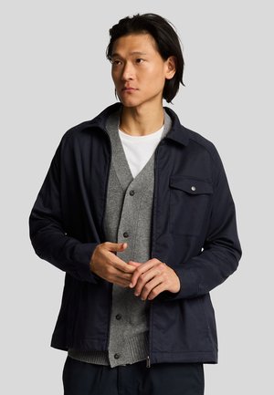 Veste navy avec une poche à boutons, coupe décontractée et fermeture éclair. Superposée sur un pull en V gris. Texture lisse, design classique.
