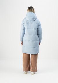 Persoon die met de rug naar de camera staat, gekleed in een lichtblauwe gewatteerde jas met capuchon, bruine wijdvallende broek en witte sneakers, tegen een effen achtergrond.