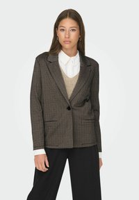 Black and brown checkered blazer with a single button, tailored fit, and flapped pockets, worn over a white shirt and beige knit sweater.