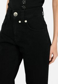 Jeans in denim nero con vita alta, caratterizzati da una chiusura con bottone argentato e dettagli decorativi in perle sulla cintura. Design classico a cinque tasche.