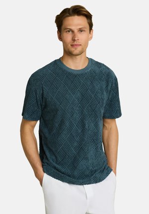Man with light brown hair wearing a textured dark teal short-sleeve shirt and white pants with hands in pockets.