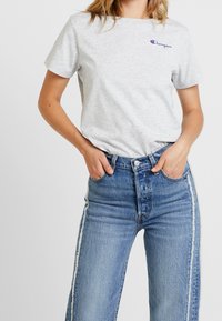 Grå kortærmet t-shirt med blåt "Champion"-logo, kombineret med højtaljede blå cowboybukser med hvide striber på siden.