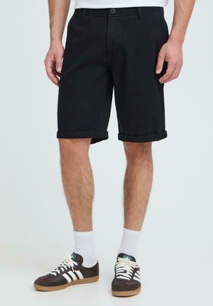 Homme portant un short noir retroussé, des chaussettes blanches et des baskets Adidas noires avec des bandes blanches, debout devant un fond uni.