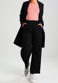 Svart skräddarsydd lång kappa, topp med rosa prickar och svarta vida byxor. Lätta skor fulländar outfiten och visar upp en modern stil.