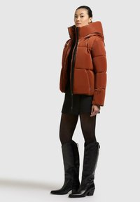 Giacca imbottita arancione con colletto alto e chiusura con zip, indossata con un mini abito nero e stivali neri al ginocchio in pelle.