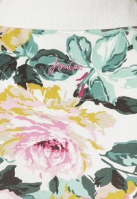 Tissu à motifs floraux avec des nuances de rose, jaune et vert sur un fond texturé. Comprend le logo "Joules" brodé en rose.