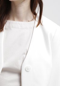 Vit blazer med en slät textur, med en enda rund knappstängning, bärs över en vit topp med en enkel, strukturerad halsringning.