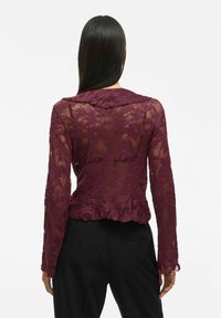 Top en dentelle bordeaux avec un ourlet peplum, des manches longues et un motif floral. La vue arrière montre un tissu texturé et un col rond. Pantalons noirs.