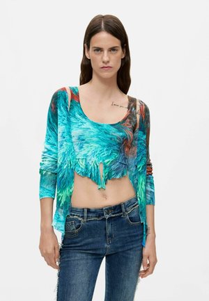 Kobieta z długimi ciemnymi włosami nosi niebiesko-zielony krótki top tie-dye z pasującym kardiganem i niebieskie dżinsy, stoi na białym tle.