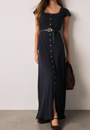 Femme portant une longue robe noire boutonnée avec manches courtes et ceinture, tenant un sac imprimé léopard et portant des chaussures assorties imprimé léopard.