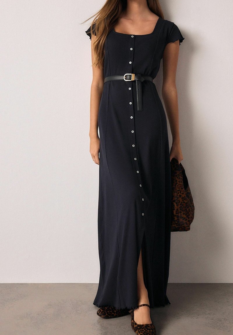 Femme portant une longue robe noire boutonnée avec manches courtes et ceinture, tenant un sac imprimé léopard et portant des chaussures assorties imprimé léopard.