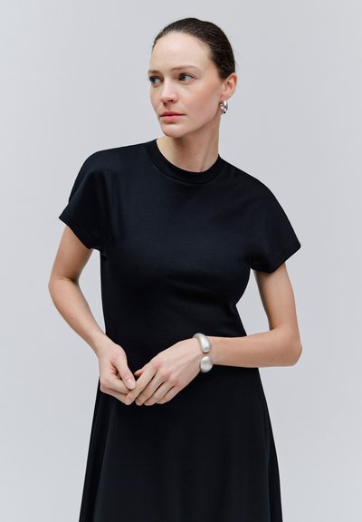 Femme en robe noire à manches courtes avec bracelet manchette argenté et boucles d'oreilles, regardant vers sa gauche les mains jointes devant elle.