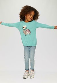 Turquoise langemouwen top met een uilontwerp van pailletten; gecombineerd met lichtblauwe jeans en grijs/roze sneakers.