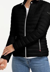 Woman wearing a black quilted jacket with silver zippers and blue jeans, adjusting sleeve over white shirt.