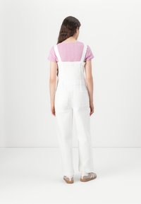 Pantalones de mezclilla blancos con tirantes ajustables, corte suelto y dos bolsillos traseros, combinados con una blusa de manga corta de color púrpura claro.