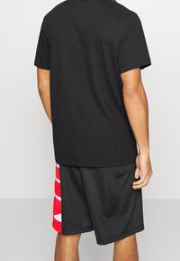 Camiseta de algodón negra con mangas cortas combinada con pantalones cortos negros que presentan un patrón de olas en rojo y blanco en el lado izquierdo. Fondo liso.