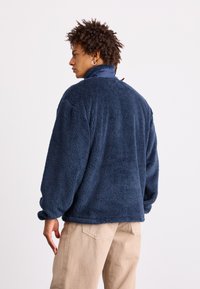 Tamno plavi flis jakna s visokim ovratnikom i dugim rukavima, koja se odlikuje mekim, teksturiranim materijalom i opuštenim krojem.