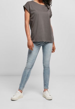 Woman wearing a loose dark gray short-sleeve shirt, light blue skinny jeans, and white sneakers standing against a plain background.