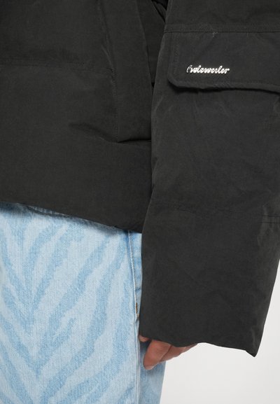 Nærbillede af en person iført en sort jakke med synlig "Woolrich"-logo på lommen på ærmet og lyseblå mønstrede denimjeans.