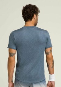 Camiseta atlética azul de tela suave, con cuello redondo clásico, mangas cortas y dobladillo recto; llevada por una persona con tatuajes.