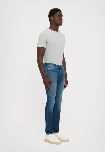 Homme portant un t-shirt gris clair, un jean bleu slim et des baskets blanches sur fond blanc uni.