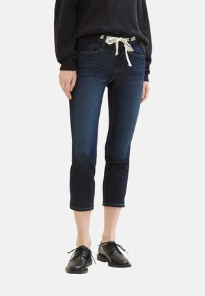 Person wearing dark cropped jeans with white drawstring belt, black sweater, and black lace-up shoes standing on white background.