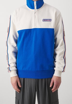 Person trägt ein weiß-blaues Halbreißverschluss-Sweatshirt mit Napapijri-Logo und gestreiften Details an den Ärmeln, kombiniert mit grauen Nadelstreifenhosen.