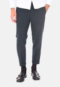 Pantaloni verde scuro su misura con una texture liscia, taglio affusolato, orli arrotolati e una leggera lucentezza, abbinati a mocassini neri.