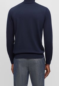 Homme portant un pull bleu marine à manches longues et un pantalon texturé gris, vu de dos sur un fond gris uni.