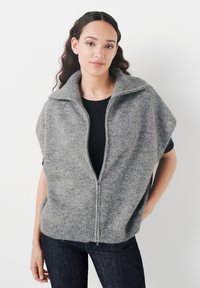 Grå zip-up väst gjord av mjukt tyg med hög krage och breda ärmhål, med ribbade fålldetaljer. Bärs över en svart topp.