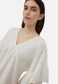 Blusa de lino blanca con un escote en V profundo y mangas cortas con volantes. La tela tiene un acabado texturizado, creando una apariencia ligera y aireada.