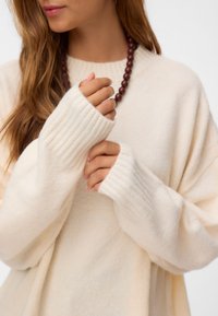 Kobieta nosząca kremowy, oversize, prążkowany sweter, trzymająca rękawy jedną ręką, nosząca ciemny naszyjnik z koralików, widoczna od ramion do ust.