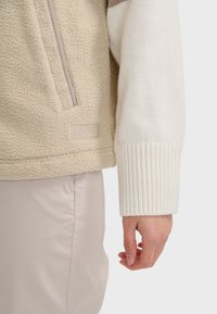 Pullover in pile color beige chiaro con finitura testurizzata, dotato di tasca con zip e maniche bianche a costine; dettaglio del logo sull'orlo.