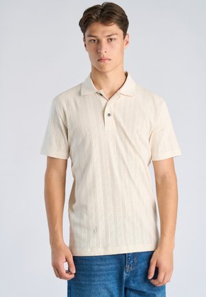 Młody mężczyzna stojący, ubrany w kremową, teksturowaną koszulkę polo z krótkim rękawem i niebieskie dżinsy na jednolitym jasnym tle.