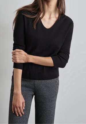 Schwarzer Strickpullover mit V-Ausschnitt und 3/4-Ärmeln, kombiniert mit grauen strukturierten Hosen, die seitliche Knopfdetails aufweisen.