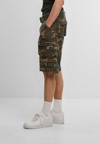 Tarnfarbenen Cargo-Shorts in Grün und Braun mit mehreren Taschen, einem Kordelzugsaum und einem Text-Logo-Patch auf der Seite. Getragen mit weißen Sneakers.