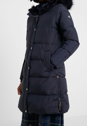 Manteau d'hiver bleu marine avec capuche en fausse fourrure, fermetures éclair et pression, poches latérales et texture matelassée. Accentué par des éléments en tan.