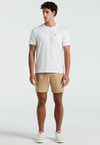 Witte katoenen t-shirt met korte mouwen en een borstzak, gecombineerd met beige linnen shorts en witte sneakers, staande tegen een lichte achtergrond.