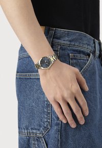 Metallisk armbåndsur med en rund marineblå urskive, gyldne detaljer og et bånd i tofarvet rustfrit stål. Båret på en hånd imod denimstof.