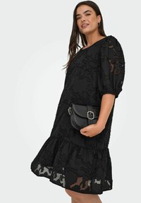 Melnas kleitas ar teksturētu ziedu rakstu, īsām pūkām un pakāpenisku apakšmalu. Apvienota ar mazu melnu quiltēto crossbody somu.