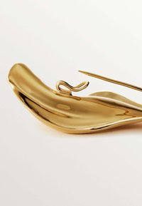 Plato de oro con forma curva, que presenta un pequeño bucle decorativo y un pincho alargado y puntiagudo al lado. Superficie lisa y reflectante.
