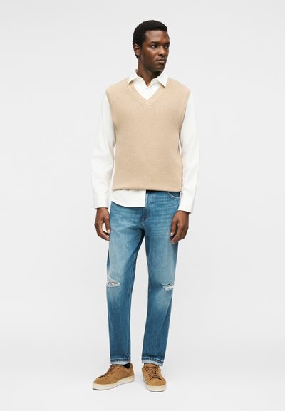 Άνδρας που φοράει μπεζ πλεκτό αμάνικο πουλόβερ πάνω από λευκό πουκάμισο με μπλε τζιν με σκισίματα και καφέ αθλητικά παπούτσια, στέκεται και κοιτάζει προς τα δεξιά.